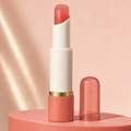 Open Trinidy MK Satin Petal Tinted Lip Balm tube displayed in a rosy-coral studio setting